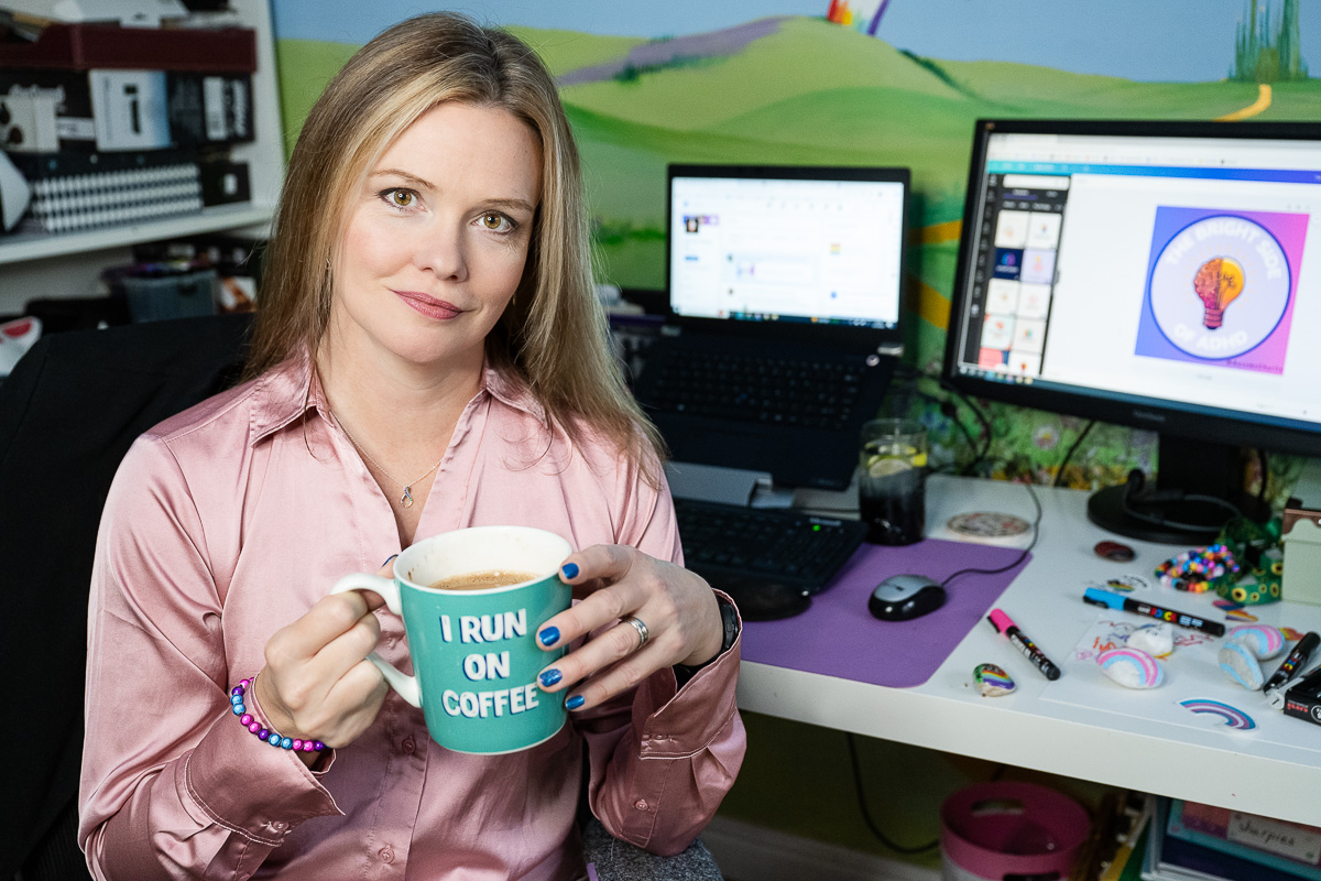 Charlie with hair and make-up done, wearing a pink sating blouse, holding a large mug of coffee with the slogan I run on coffee, desk behind her with laptop showing LinkedIn and monitor showing a Canva design