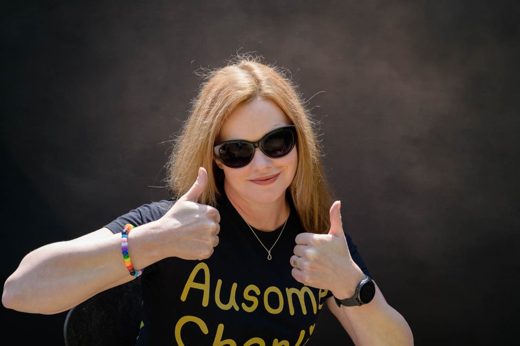 Charlie wearing dark sunglasses, progress pride bracelet, black t-shirt with Ausome Charlie in big gold letters, smiling awkwardly and holding up both thumbs.
