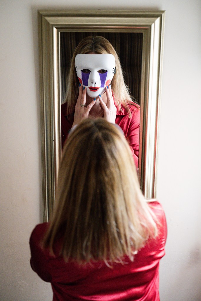 Photo of Charlie from behind, wearing a red satin shirt, her shoulder-length dark blonde highlighted hair straightened, looking in gold framed full length mirror wearing a white mask painted like clown-like with red lips and cheeks and purple tears below the eyes (a character from Five Nights at Freddy's)