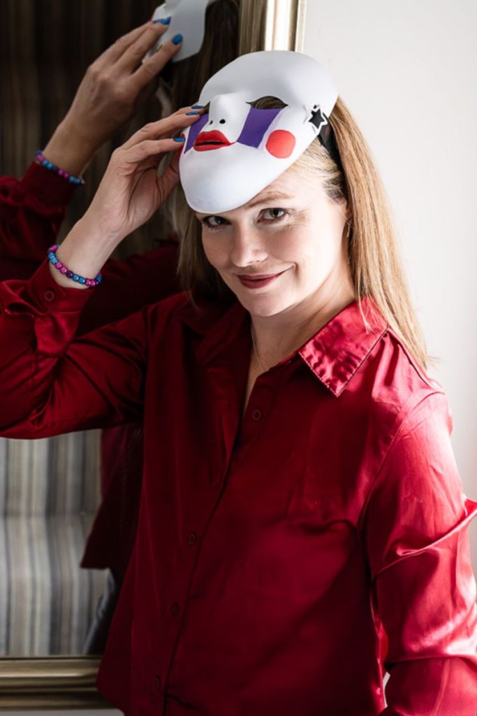 Charlie wearing a red satin shirt, raising to the top of her head a papier maché mask she had been wearing, which is painted with a sad looking character with large purple tears streaking from the eyes (a character from Five Nights at Freddie's). Underneath the mask she is smiling a close-lipped but genuine smile.