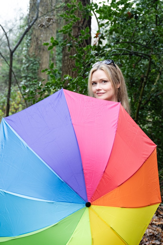 Charlie shyly peeking out over the top of a large rainbow umbrella which is open and held in front of her