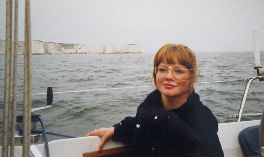 Charlie as a 15-year-old with golden blonde hair with a fringe, pink lipstick, and large round gold framed glasses, smiling awkwardly on a boat in Studland Bay, Dorset.