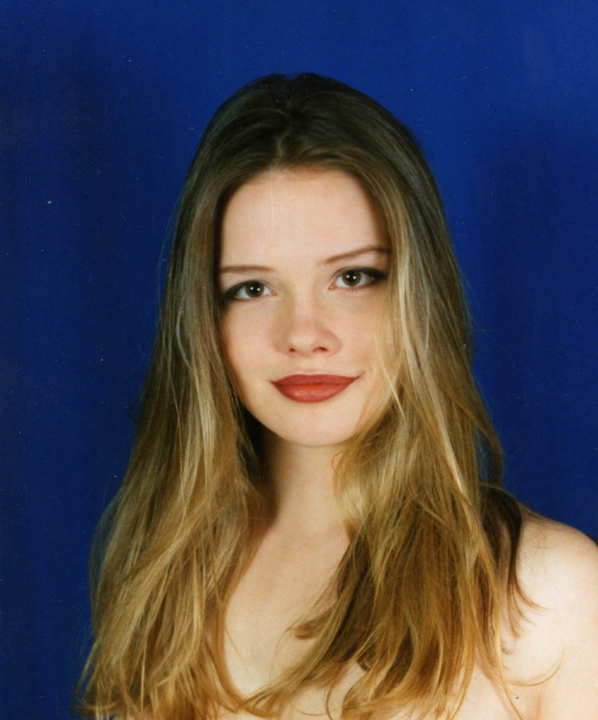 Head and shoulders photo of Charlie as a 20-year-old with long, blonde-highlighted hair, cherry lipstick, dark eye makeup, no glasses, no clothes, smiling awkwardly in front of a blue screen.