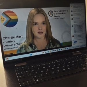 Photo of an open laptop with the screen showing Charlie speaking at a Neurodiversity Celebration Week online live zoom webinar event. Charlie is wearing a green satin shirt, her hair straightened, and her Zoom background shows Neurodiversity Celebration Week and she has added Charlie Hart she/they Ausome Charlie www/AusomeCharlie.co.uk and an image of an intersex inclusive pride progress flag button badge. 